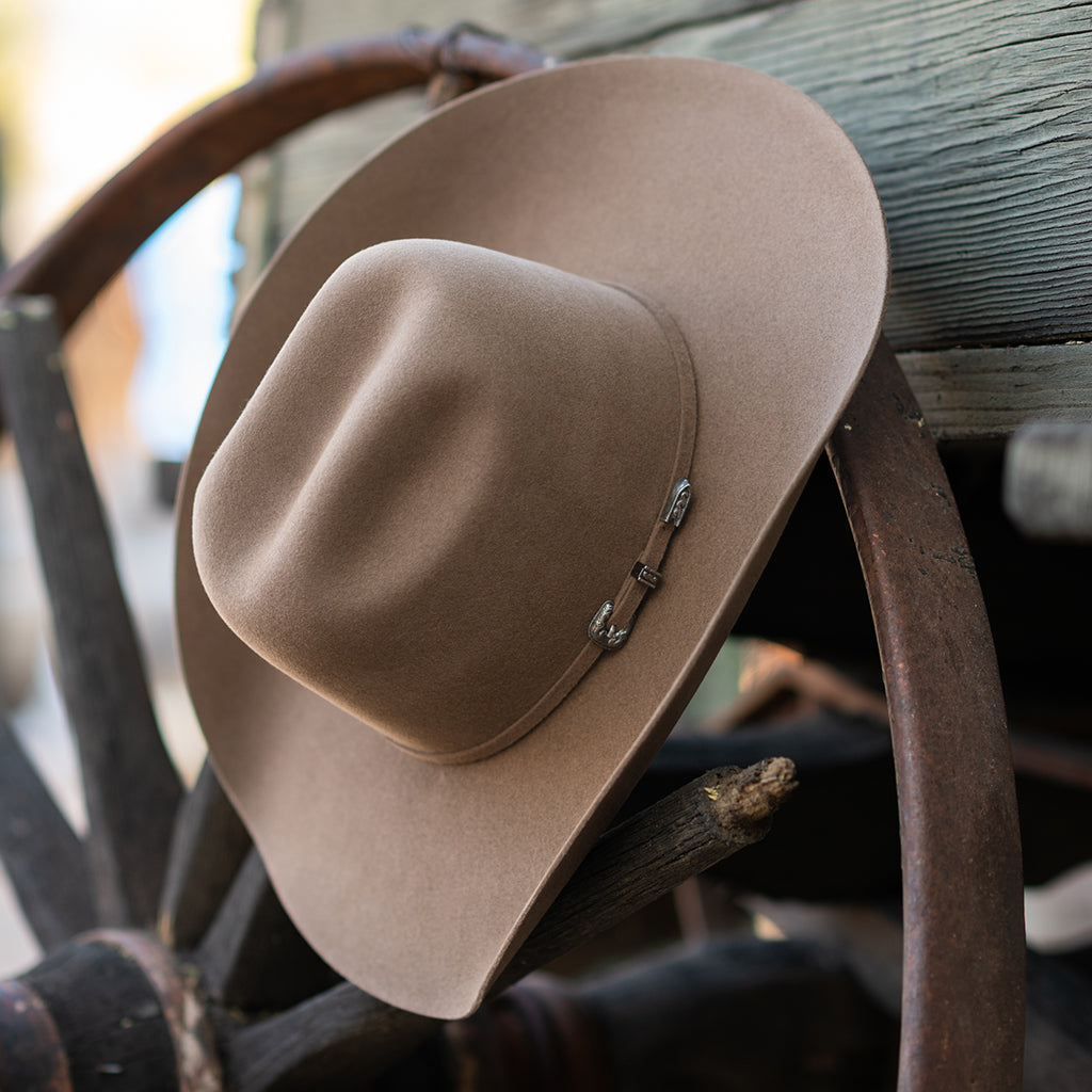 A fawn colored felt cowboy hat with silver concho-style hardware, resting on a rustic wooden wheel and plank background.