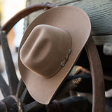 A fawn colored felt cowboy hat with silver concho-style hardware, resting on a rustic wooden wheel and plank background.