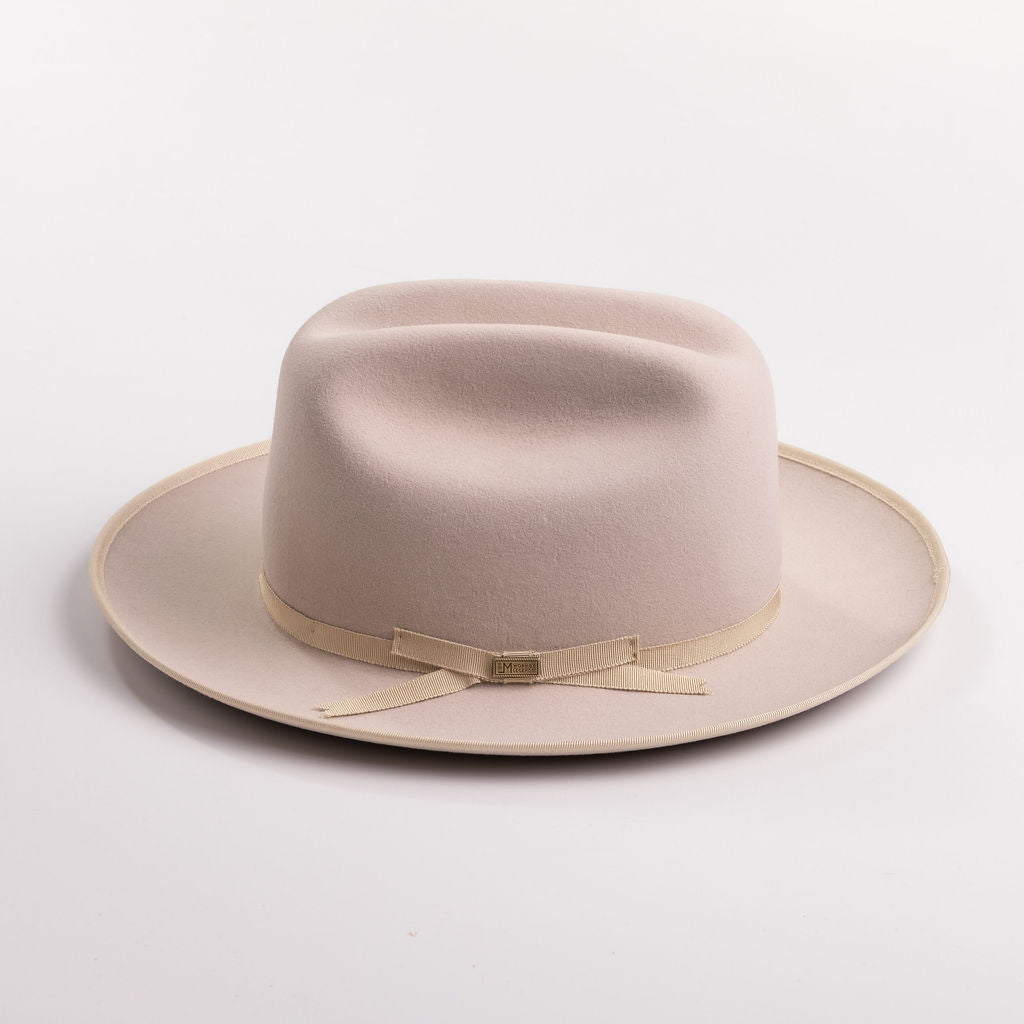 A light beige belly colored felt hat with a matching beige ribbon band and small gold-tone buckle, photographed on a light gray background