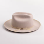 A light beige belly colored felt hat with a matching beige ribbon band and small gold-tone buckle, photographed on a light gray background