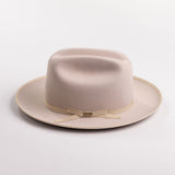 A light beige belly colored felt hat with a matching beige ribbon band and small gold-tone buckle, photographed on a light gray background