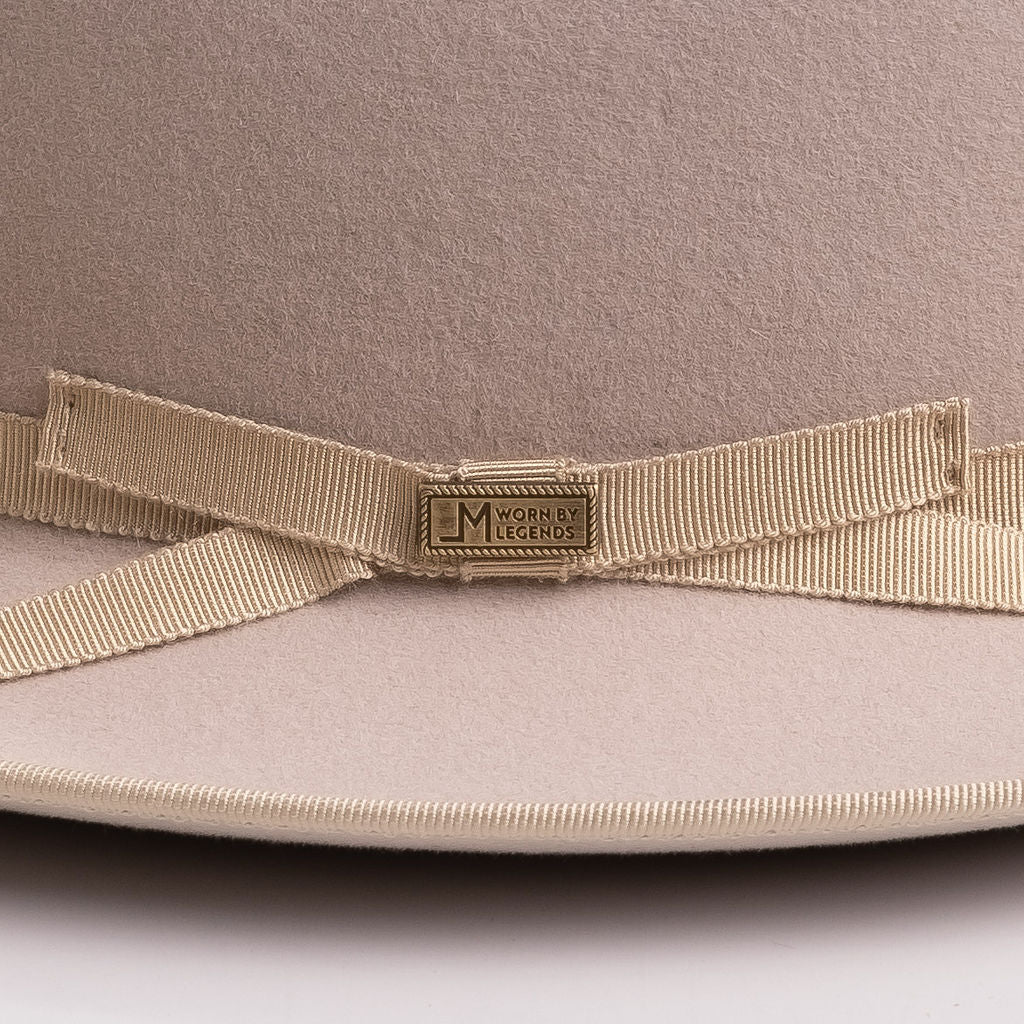 Close-up of a beige felt hat with a matching grosgrain ribbon bow and a central gold-tone metal plaque engraved with the brand "LM WORN BY LEGENDS"