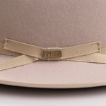 Close-up of a beige felt hat with a matching grosgrain ribbon bow and a central gold-tone metal plaque engraved with the brand "LM WORN BY LEGENDS"