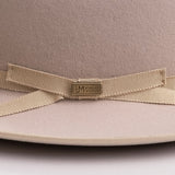 Close-up of a beige felt hat with a matching grosgrain ribbon bow and a central gold-tone metal plaque engraved with the brand "LM WORN BY LEGENDS"