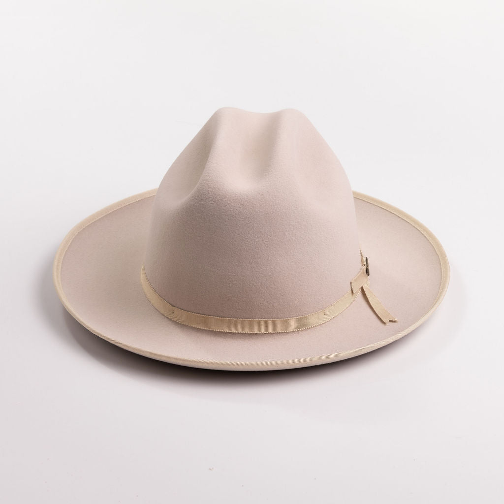 A light beige belly colored felt hat with a matching beige ribbon band and small gold-tone buckle, photographed on a light gray background