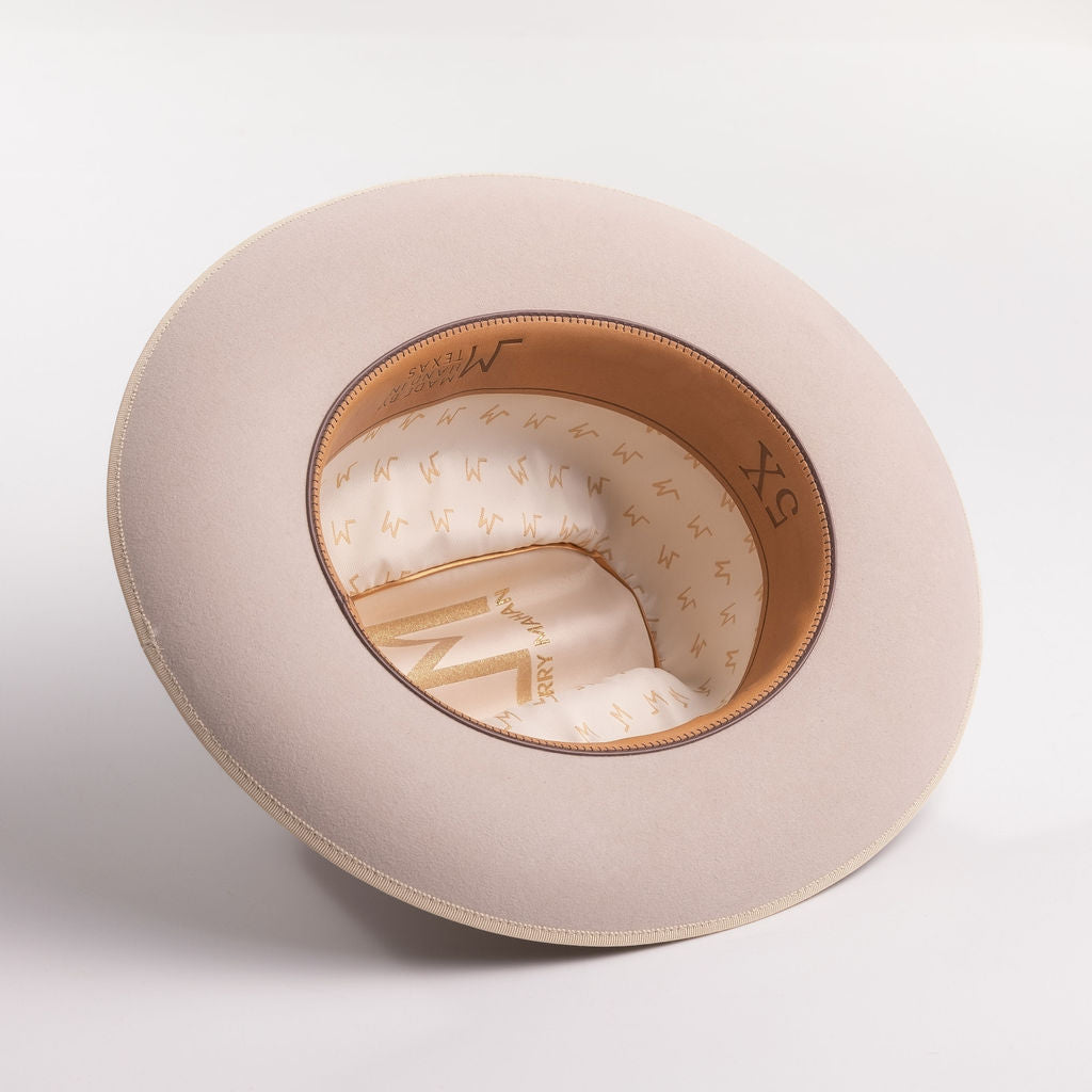 A light beige belly colored wide-brim hat shown from an overhead angle, displaying its tan leather trim and cream-colored interior patterned with the brand's logo and 5X text