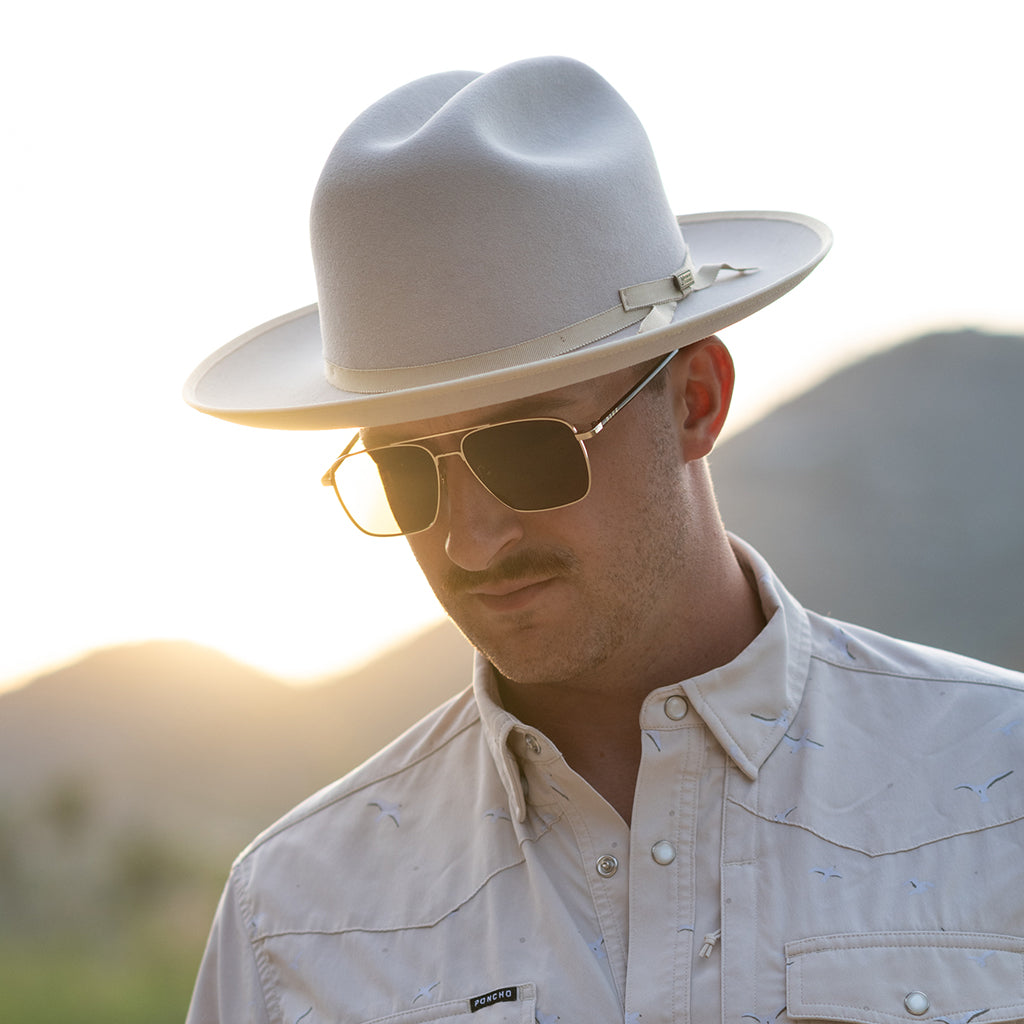 A man wearing a belly colored wide-brim fedora hat, gold-framed sunglasses, and a matching light beige western-style button-up shirt, photographed outdoors with a blurred mountain background at sunset.