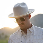 A man wearing a belly colored wide-brim fedora hat, gold-framed sunglasses, and a matching light beige western-style button-up shirt, photographed outdoors with a blurred mountain background at sunset.