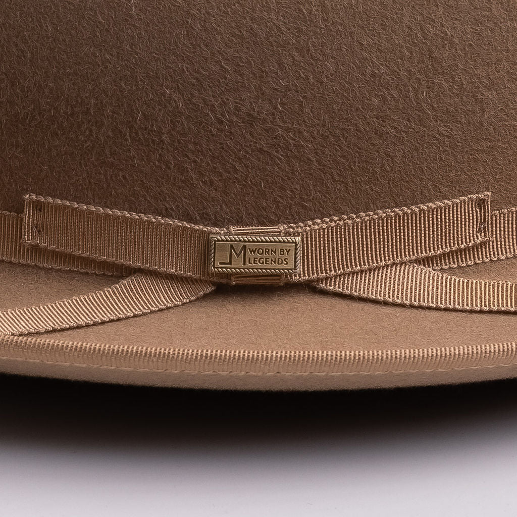 Close-up of a brown felt hat with a matching grosgrain ribbon bow and a small metal brand plaque reading "LM WORN BY LEGENDS"