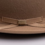 Close-up of a brown felt hat with a matching grosgrain ribbon bow and a small metal brand plaque reading "LM WORN BY LEGENDS"