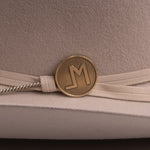 Close-up of a beige felt hat with a cream ribbon band and a round gold-tone metal emblem featuring a stylized letters "LM"