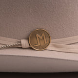 Close-up of a beige felt hat with a cream ribbon band and a round gold-tone metal emblem featuring a stylized letters "LM"