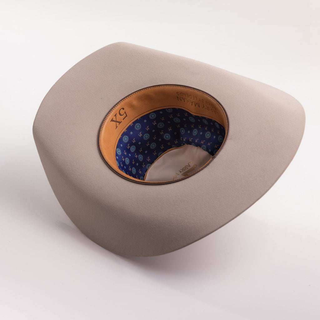 A light taupe wide-brimmed cowboy hat with a blue patterned inner lining and a tan leather-like band, photographed on a clean light gray background