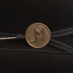 A close-up of a black hatband featuring a round bronze-colored metal medallion engraved with the letter "LM", tied with black ribbon on a black velvet background.