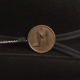 A close-up of a black hatband featuring a round bronze-colored metal medallion engraved with the letter "LM", tied with black ribbon on a black velvet background.