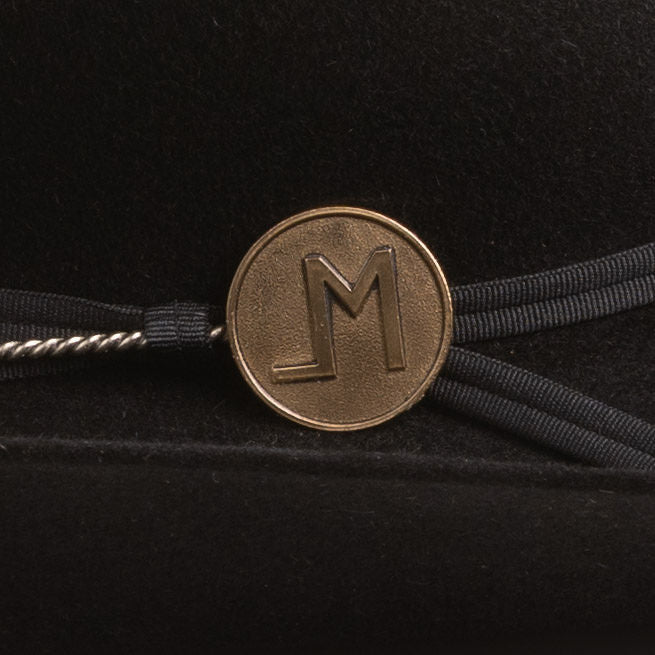 A close-up of a black hatband featuring a round bronze-colored metal medallion engraved with the letter "LM", tied with black ribbon on a black velvet background.