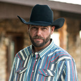 A man wearing a black cowboy hat and a blue patterned button-down shirt, photographed outdoors with a blurred natural background