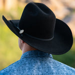 A man viewed from behind wearing a black cowboy hat with a black band and a silver metal coin with "LM", paired with a blue patterned shirt against a blurred outdoor background