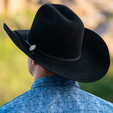 A man viewed from behind wearing a black cowboy hat with a black band and a silver metal coin with "LM", paired with a blue patterned shirt against a blurred outdoor background