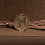A close-up of a brown ribbon headband with a round bronze-toned metal charm featuring an embossed letters "LM", resting on a textured brown surface.
