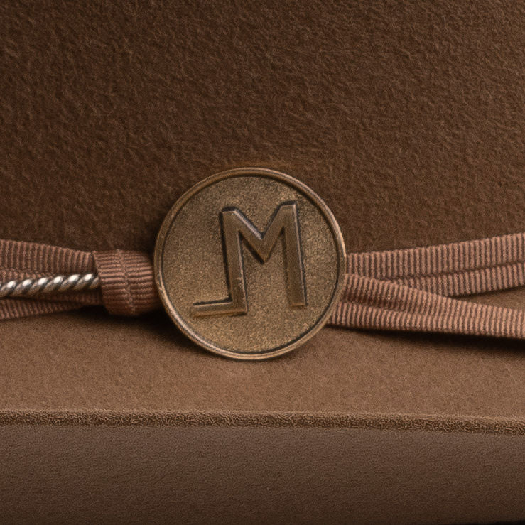 A close-up of a brown ribbon headband with a round bronze-toned metal charm featuring an embossed letters "LM", resting on a textured brown surface.