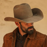 A man wearing a fawn colored cowboy hat, brown leather jacket, and dark brown shirt, looking off to the side against a warm beige background.