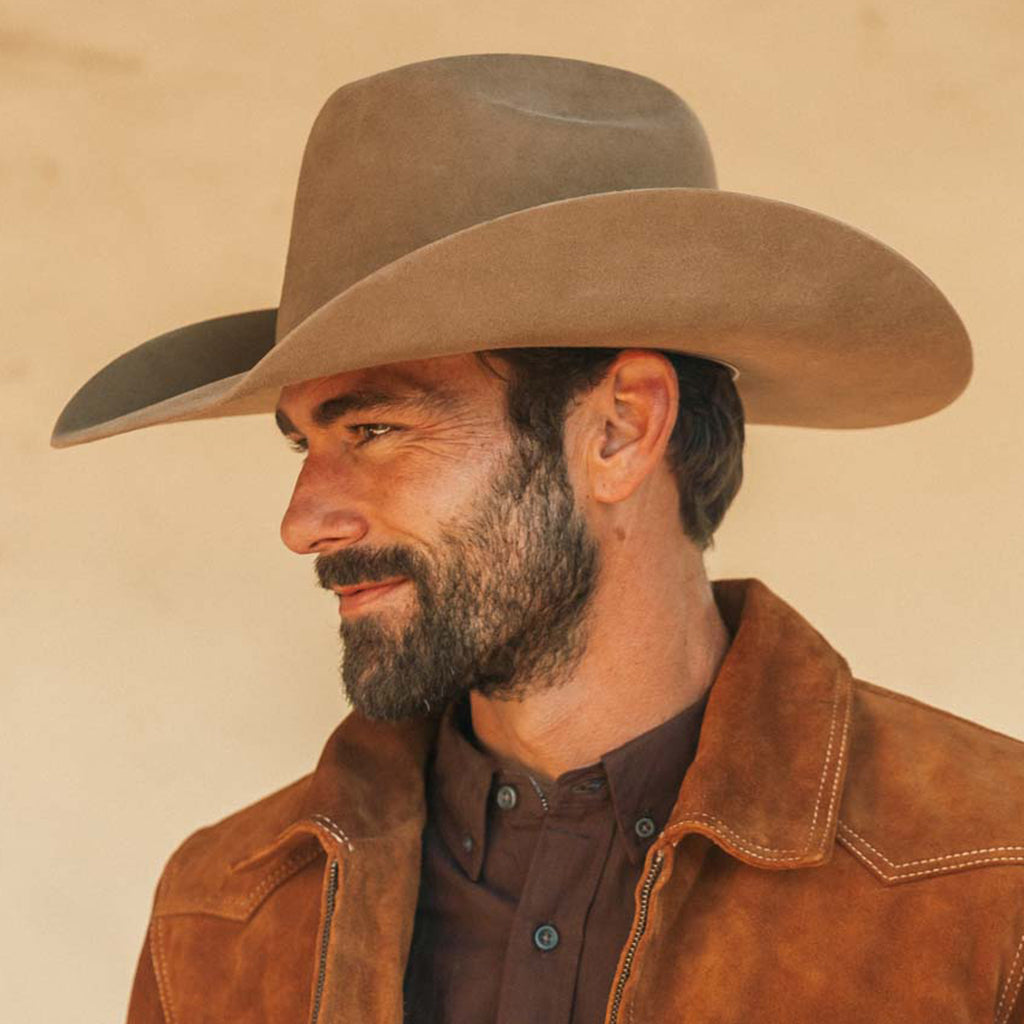 A man in profile wearing a fawn colored cowboy hat and a brown suede jacket over a dark shirt, photographed against a warm beige background