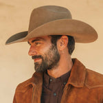 A man in profile wearing a fawn colored cowboy hat and a brown suede jacket over a dark shirt, photographed against a warm beige background