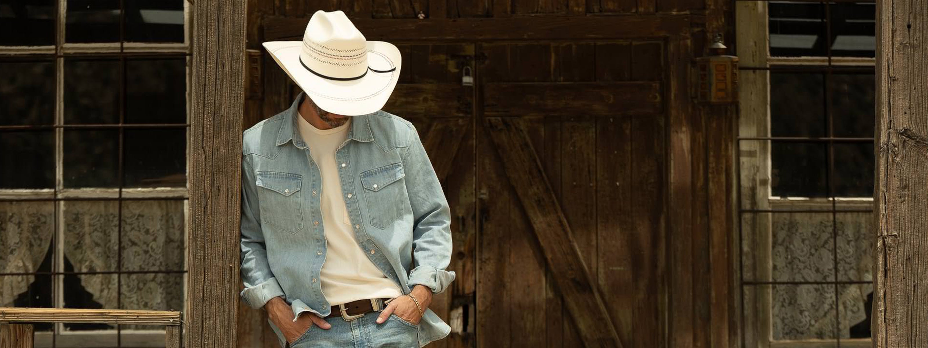 Man wearing a cowboy hat and denim jacket standing in front of a wooden door.