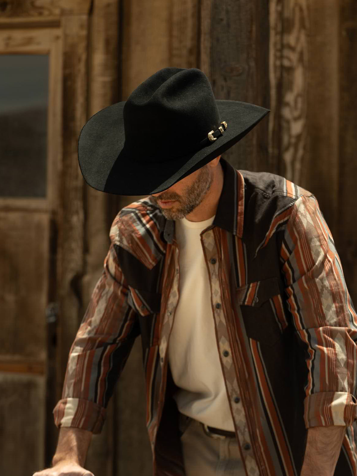 A man wearing a black cowboy hat with a small gold-toned decorative accent, a brown and beige striped button-up shirt over a white t-shirt, standing in front of a rustic wooden wall.