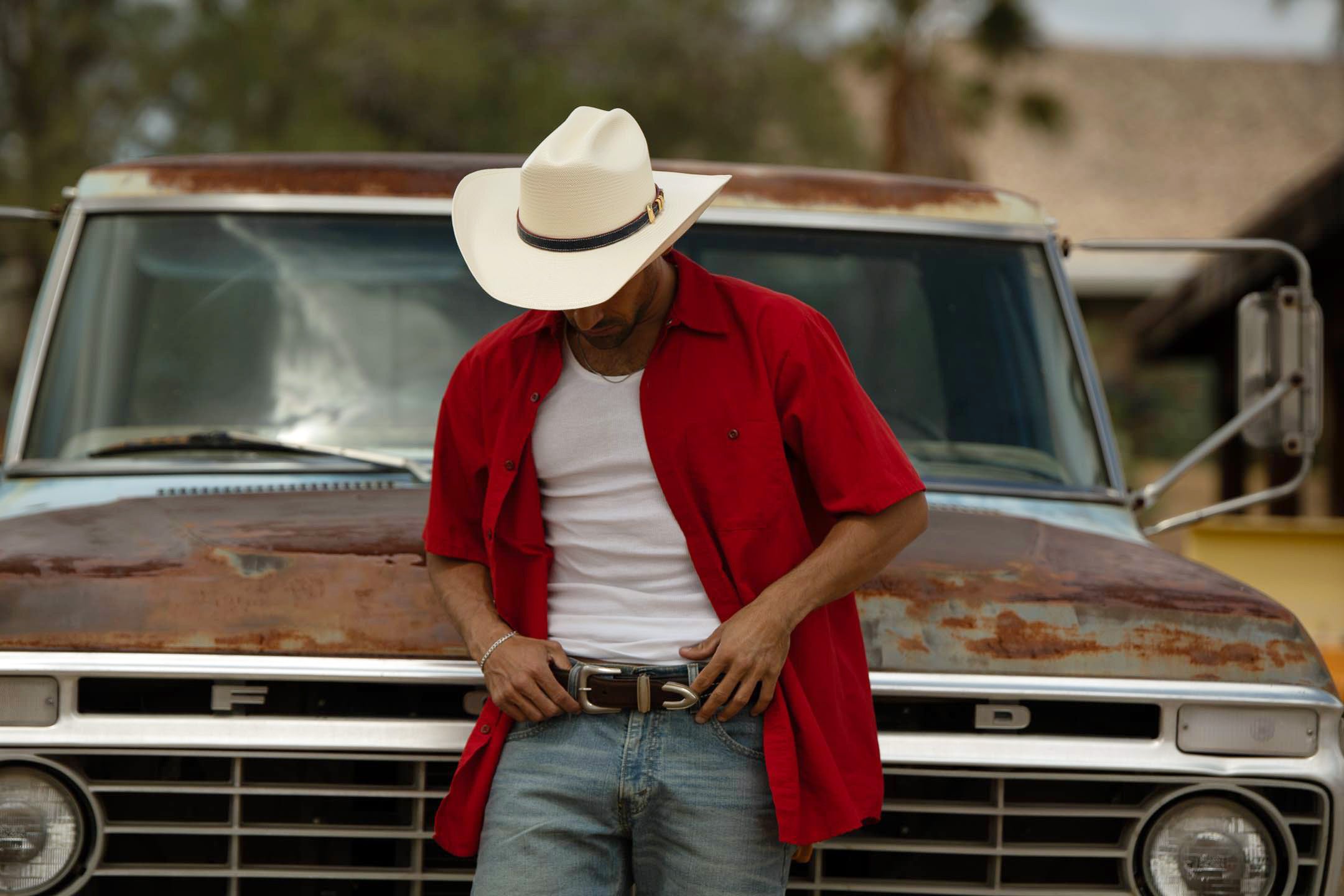 Man in a red shirt and cowboy hat standing in front of an old rusted truck.