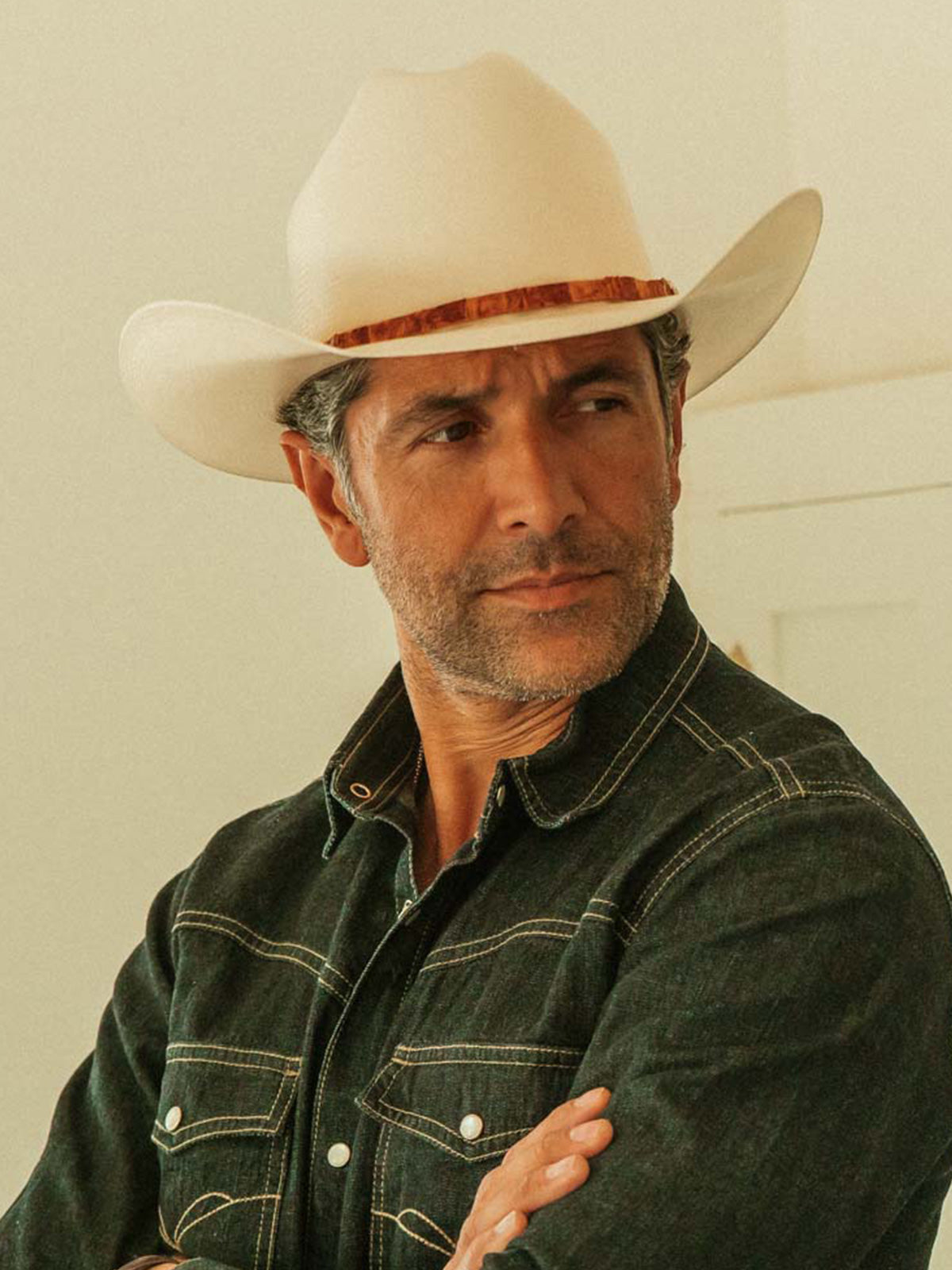 A man wearing a cream-colored cowboy hat with a buttercup band and a dark denim shirt, photographed in a brightly lit indoor setting