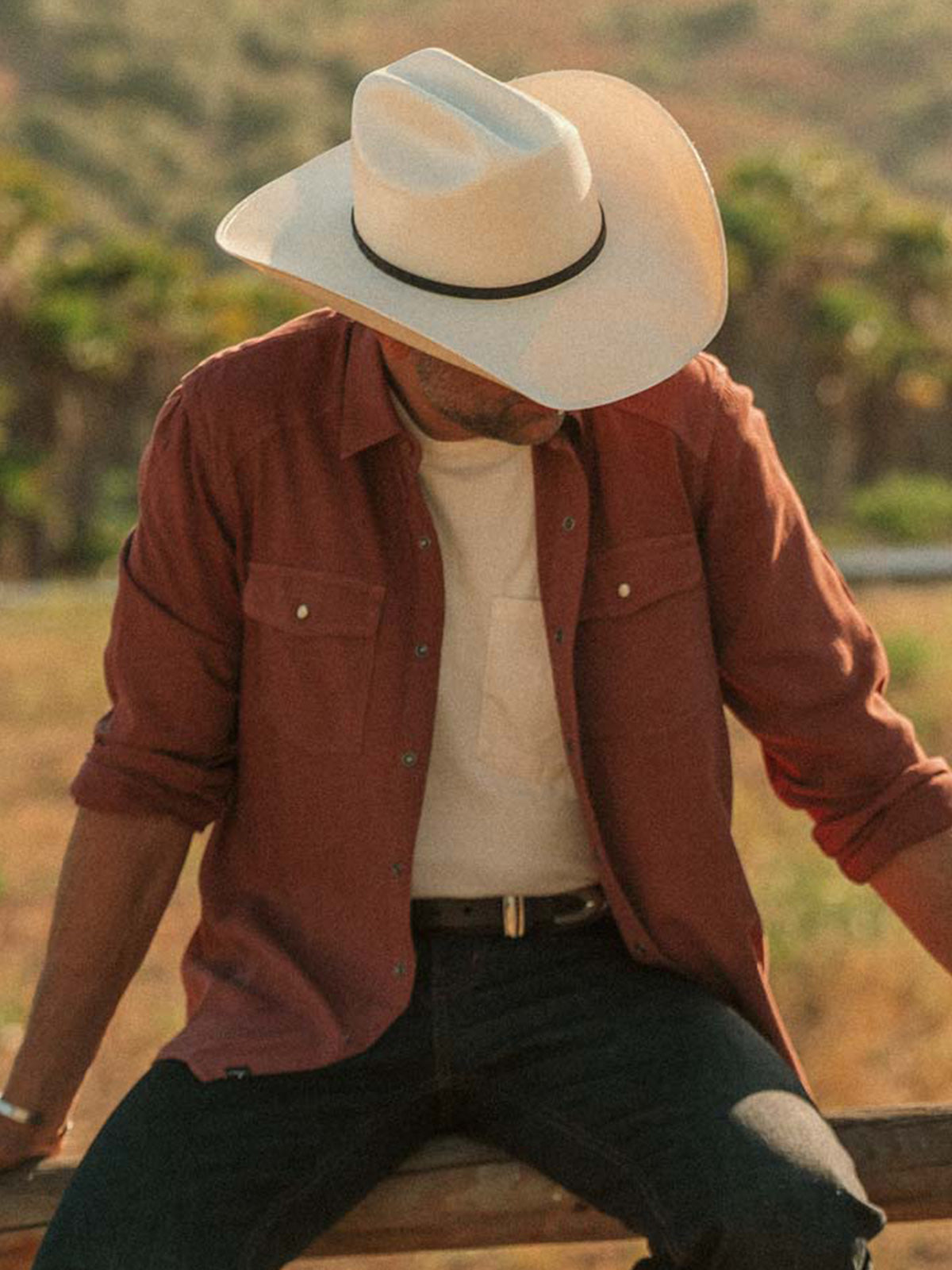 A man wearing a white cowboy hat, a rust-brown button-up shirt over a white t-shirt, and dark jeans, sitting outdoors on a wooden bench