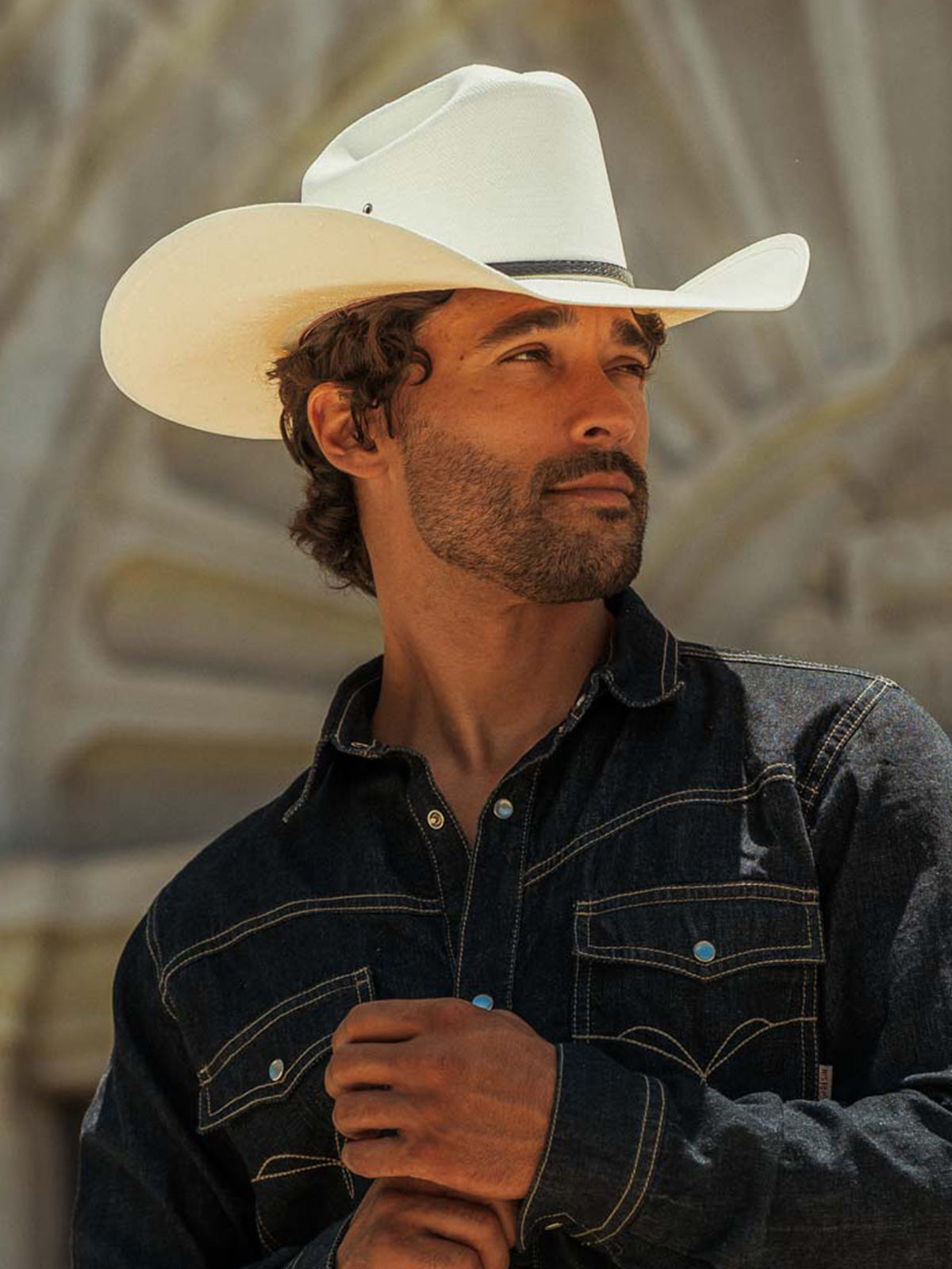 A man wearing a white cowboy hat and a dark denim shirt, standing in front of a large stone architectural structure