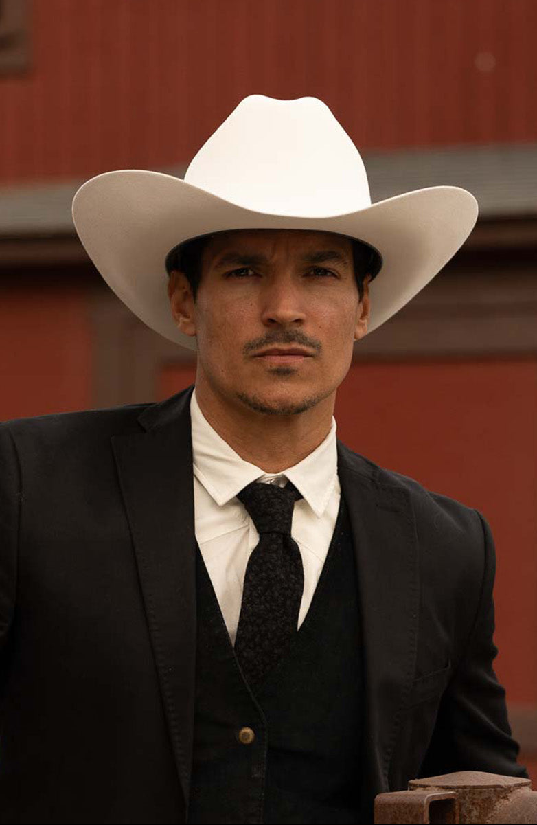 Man wearing a white cowboy hat and formal suit in front of a red barn.