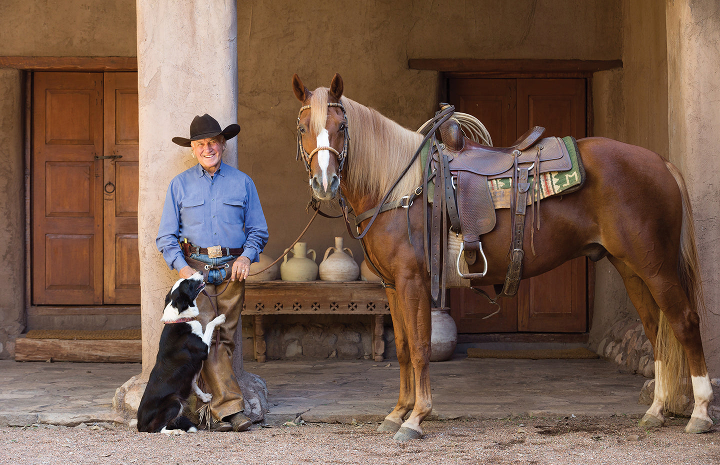 Larry Mahan wearing a brown cowboy hat, blue long sleeve shirt, and brown pants standing next to his brown horse and black and white dog while standing against a sandy cement beam