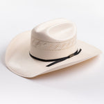 A light cream-colored woven cowboy hat with a black ribbon band and a small gold logo tag, photographed on a clean light gray background