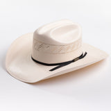A light cream-colored woven cowboy hat with a black ribbon band and a small gold logo tag, photographed on a clean light gray background