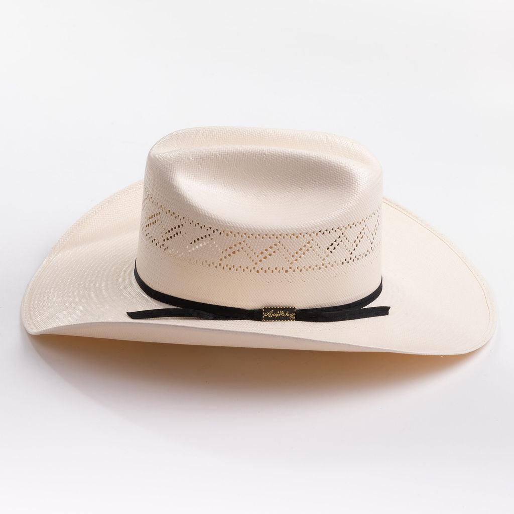 A light cream-colored woven cowboy hat with a black ribbon band and a small gold logo tag, photographed on a clean light gray background