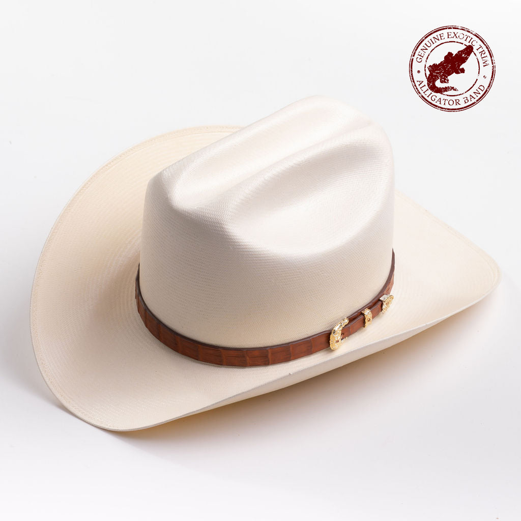 A cream-colored cowboy hat with a brown band and gold-colored decorative studs, photographed on a clean light gray background; genuine exotic trim - alligator band in red is stamped in the top right corner of the photo