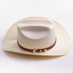 A cream-colored cowboy hat with a brown band and gold-colored decorative studs, photographed on a clean light gray background