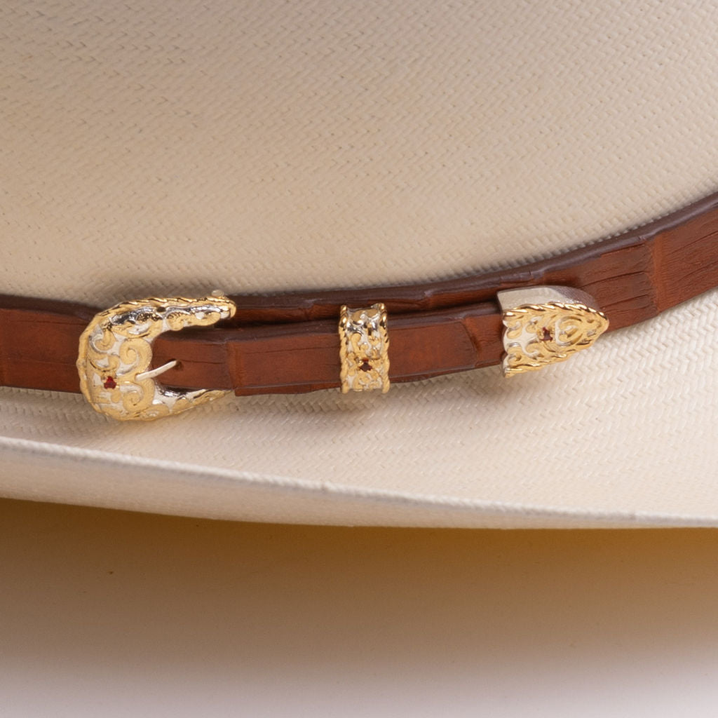 A close-up of a brown leather hat belt with ornate gold-toned concho and decorative buckle details, resting on a cream-colored cowboy hat