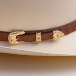 A close-up of a brown leather hat belt with ornate gold-toned concho and decorative buckle details, resting on a cream-colored cowboy hat