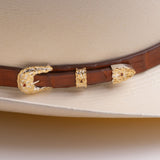 A close-up of a brown leather hat belt with ornate gold-toned concho and decorative buckle details, resting on a cream-colored cowboy hat