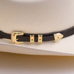 Close-up of a black textured hat band with gold-tone hardware and small red accents, shown wrapped around a cream-colored cowboy hat