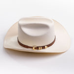 A cream-colored cowboy hat with a brown leather band and gold buckle, photographed on a clean light gray background