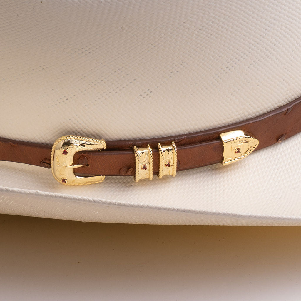 A close-up of a brown leather hat band with gold-tone metal accents and small red decorative studs, shown wrapped around a light cream-colored cowboy hat