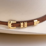 A close-up of a brown leather hat band with gold-tone metal accents and small red decorative studs, shown wrapped around a light cream-colored cowboy hat