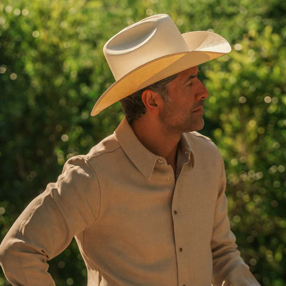 A man wearing a light beige cowboy hat and a matching beige button-up shirt, photographed outdoors against a blurred green foliage background