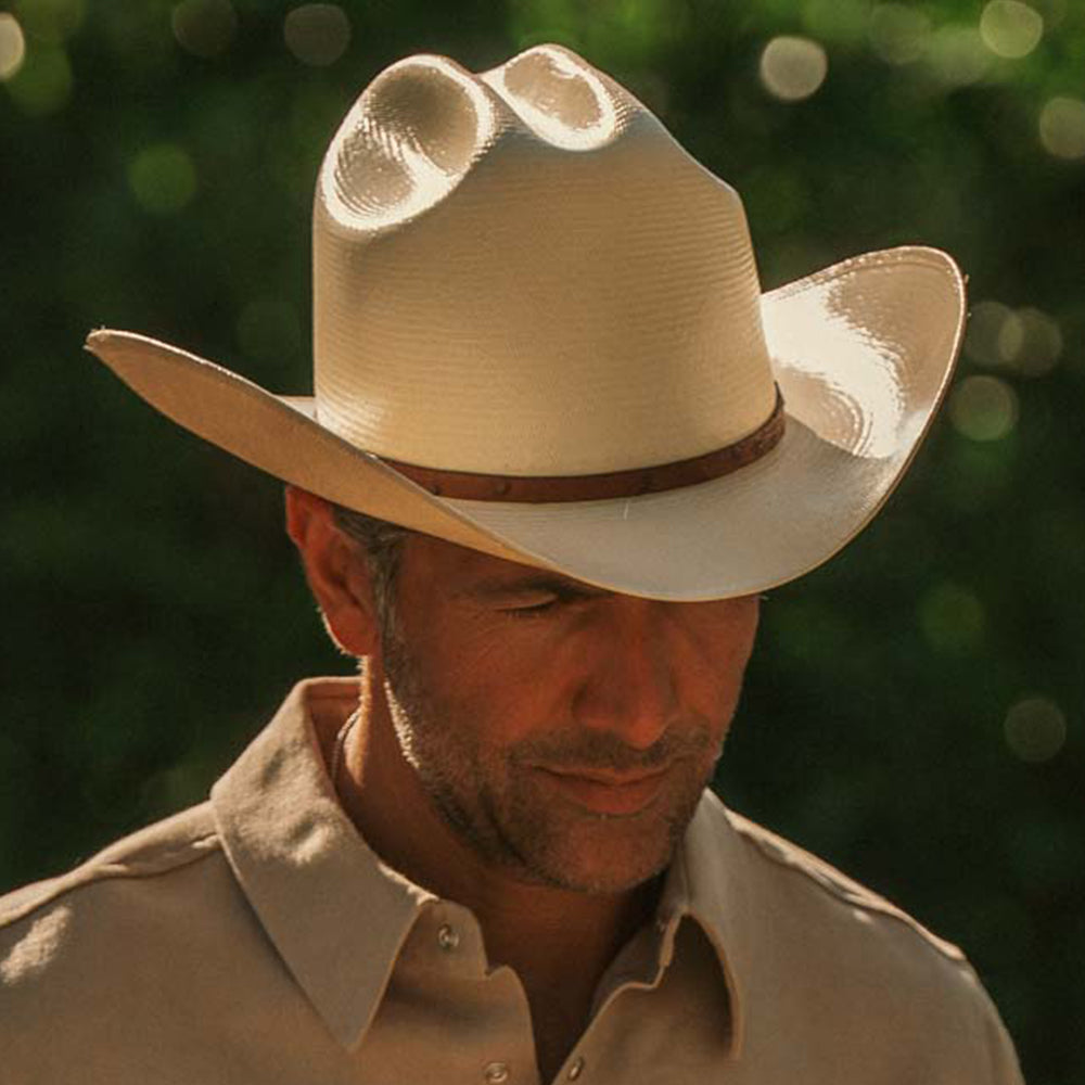 A man wearing a light beige cowboy hat with a brown band and a matching beige button-up shirt, photographed outdoors against a blurred green bokeh background
